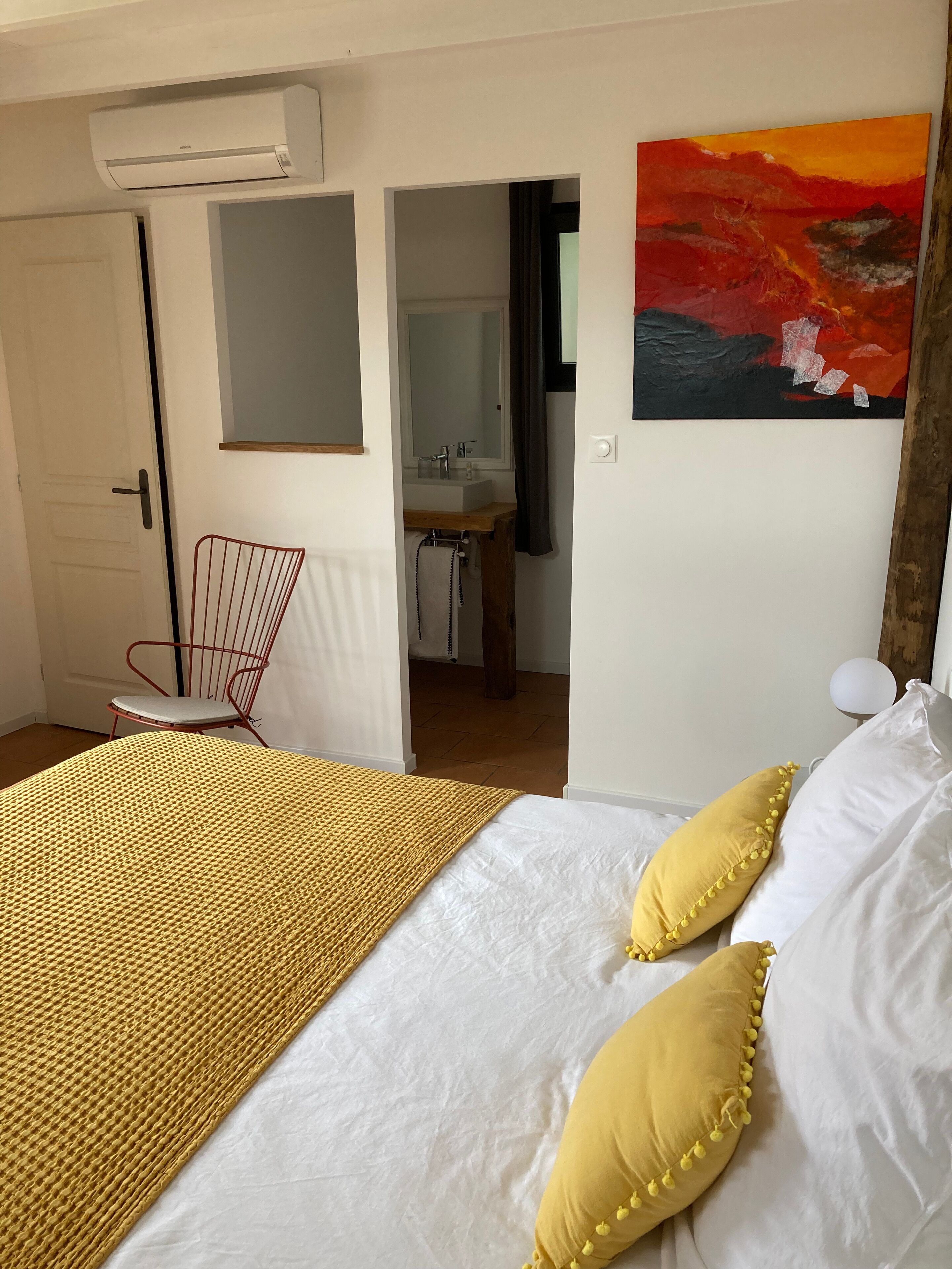 A bedroom with a bed, a chair, a painting, and a bathroom visible through an open door.