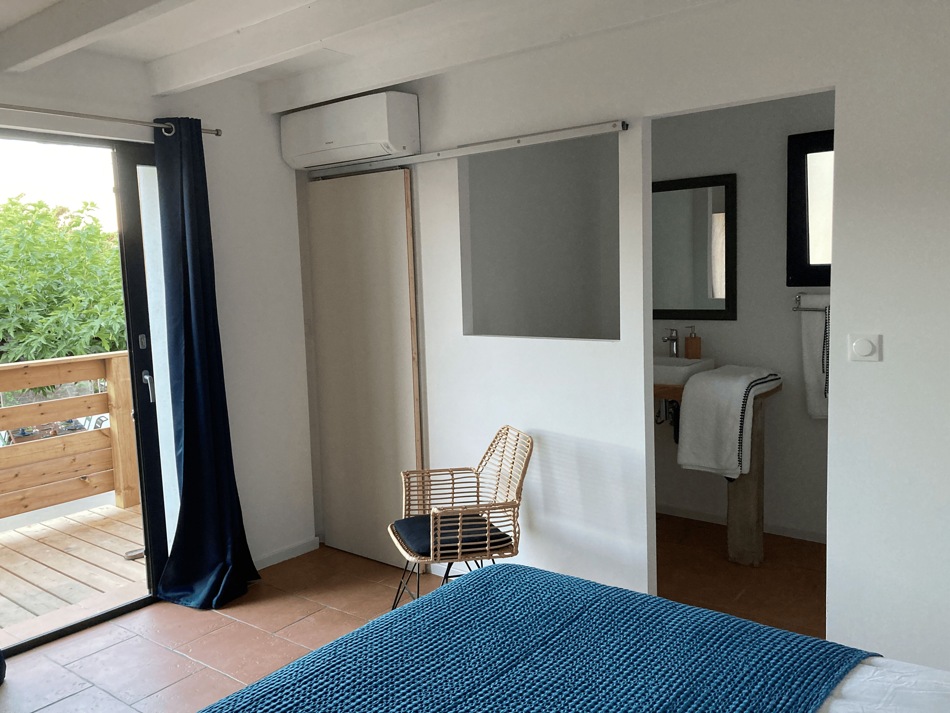 A bedroom with a bed, a chair, a bathroom with a sink and mirror, and an air conditioning unit.