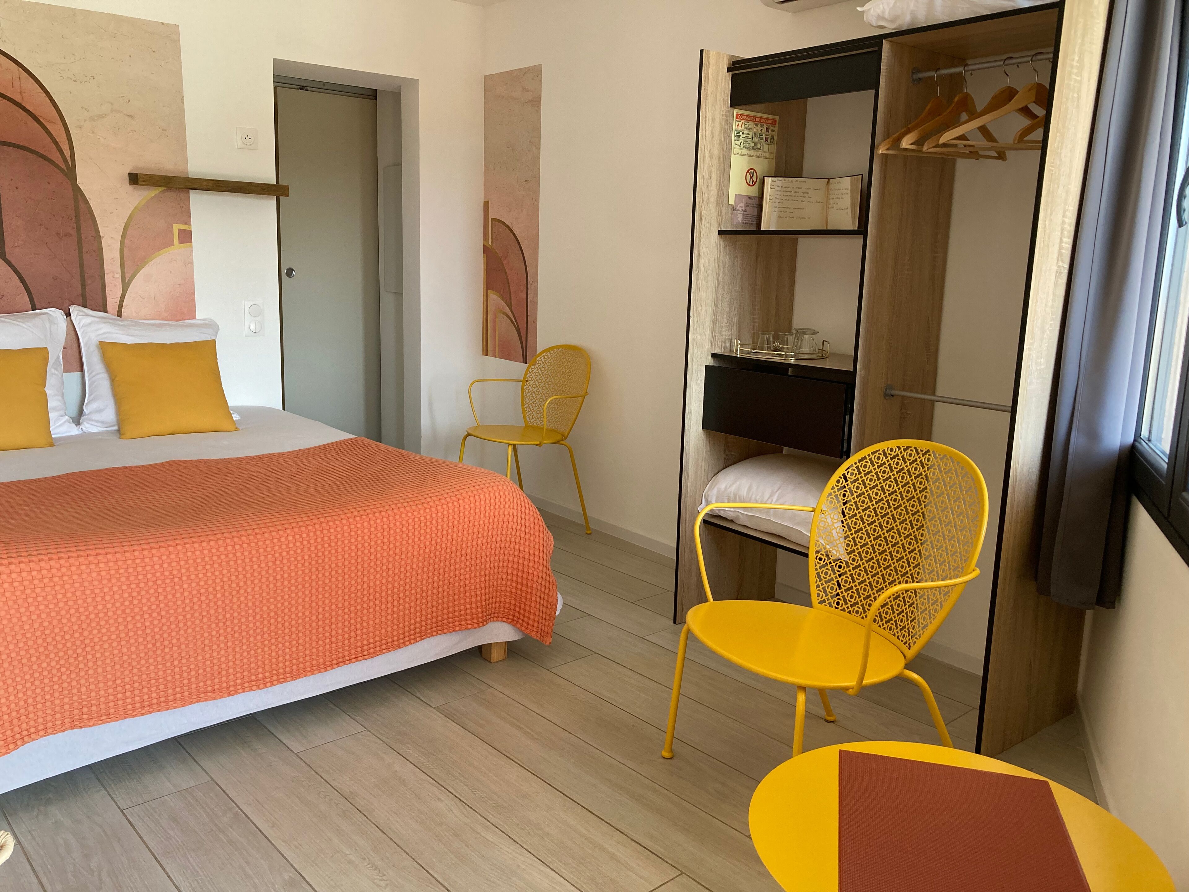 A hotel room with a bed, a yellow chair, a small table, a wardrobe, and a mirror.