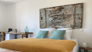 A bedroom with a bed, a bench, a table with a kettle, a vase, and a wall-mounted artwork.