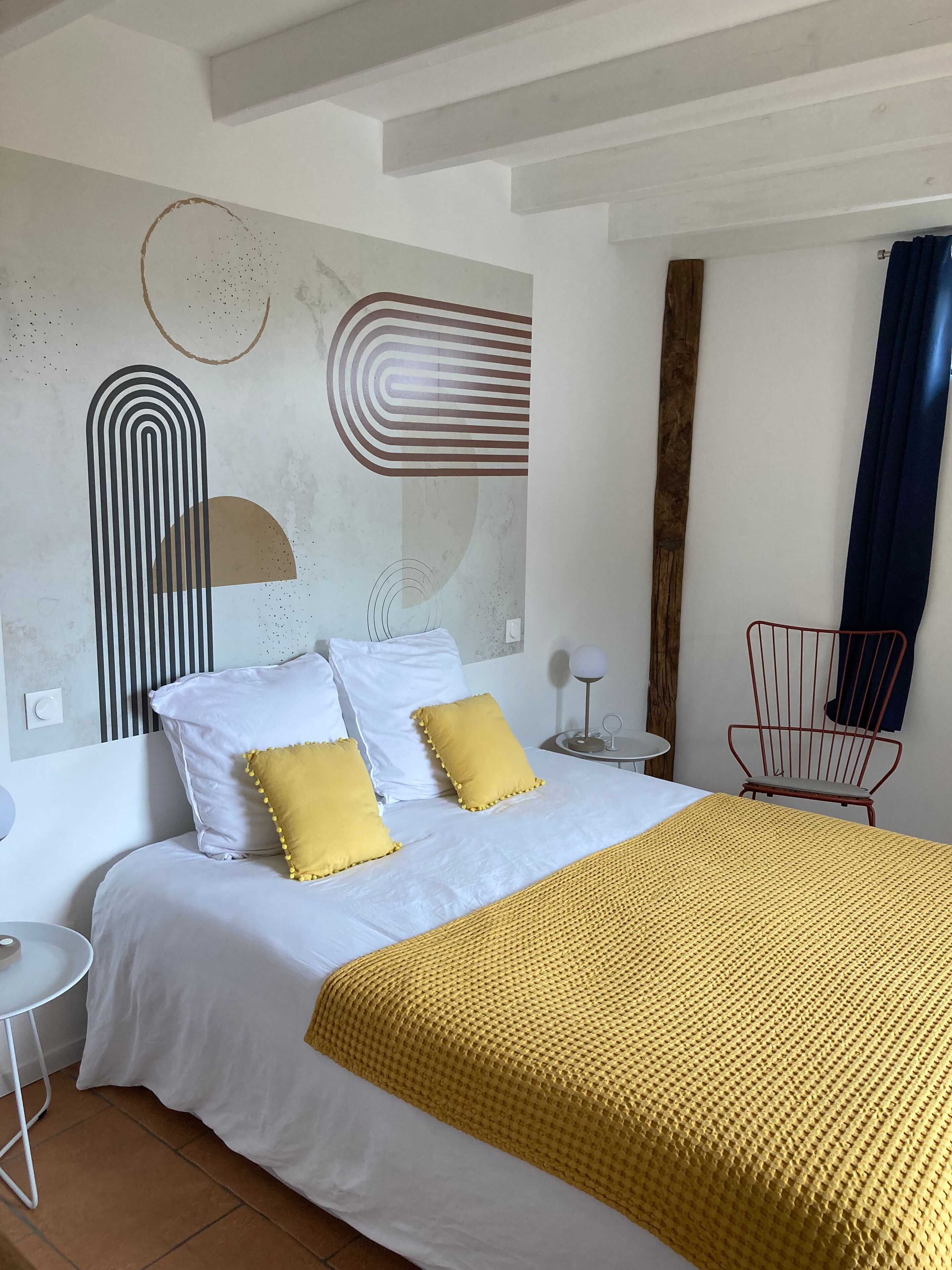 Un dormitorio moderno con una cama grande, ropa de cama blanca y amarilla, un cuadro abstracto geométrico, una pared de acento de madera y una silla roja.