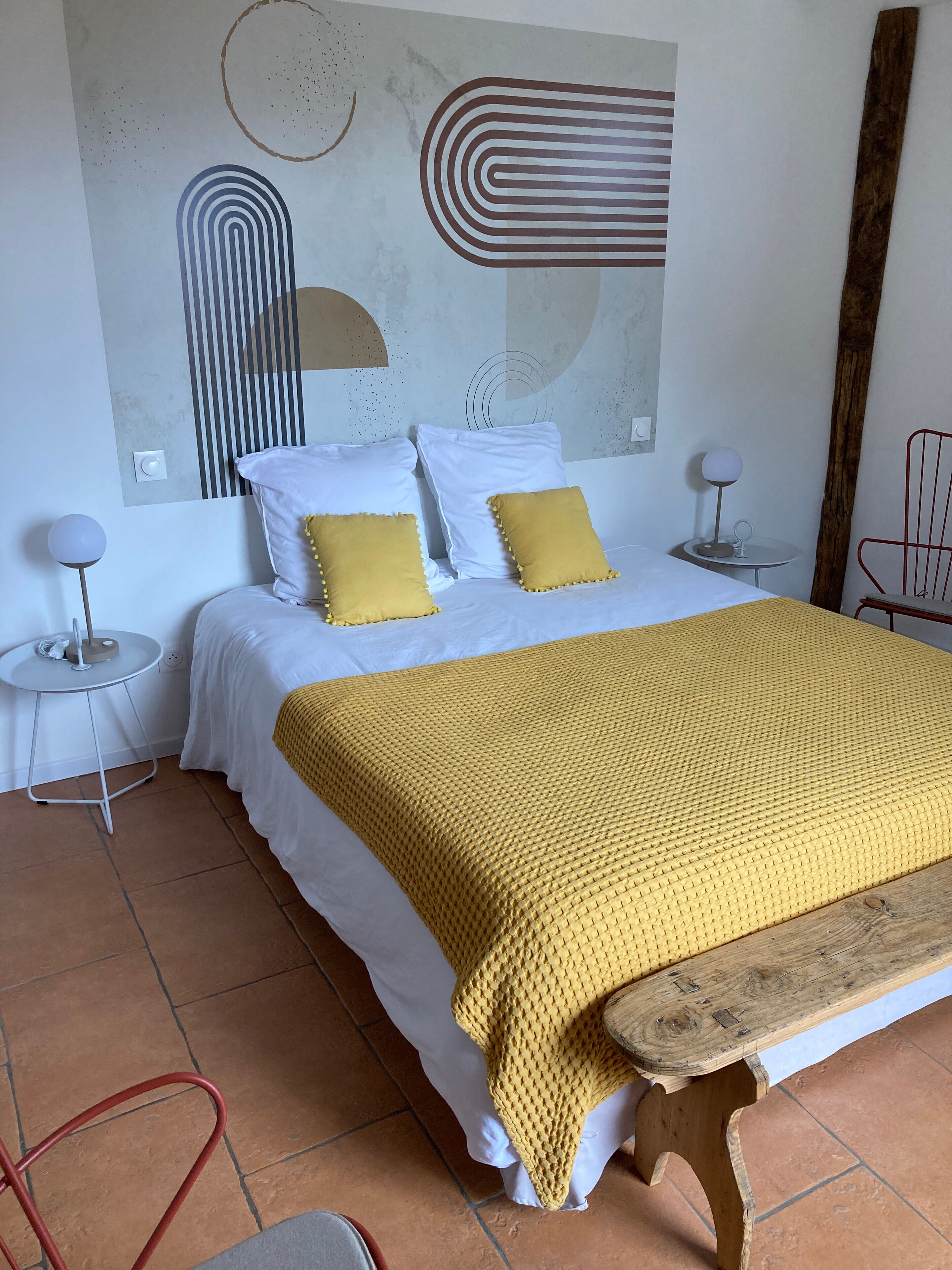 Un letto con lenzuola bianche e due cuscini gialli, una panca in legno e una parete con opere d'arte astratte.