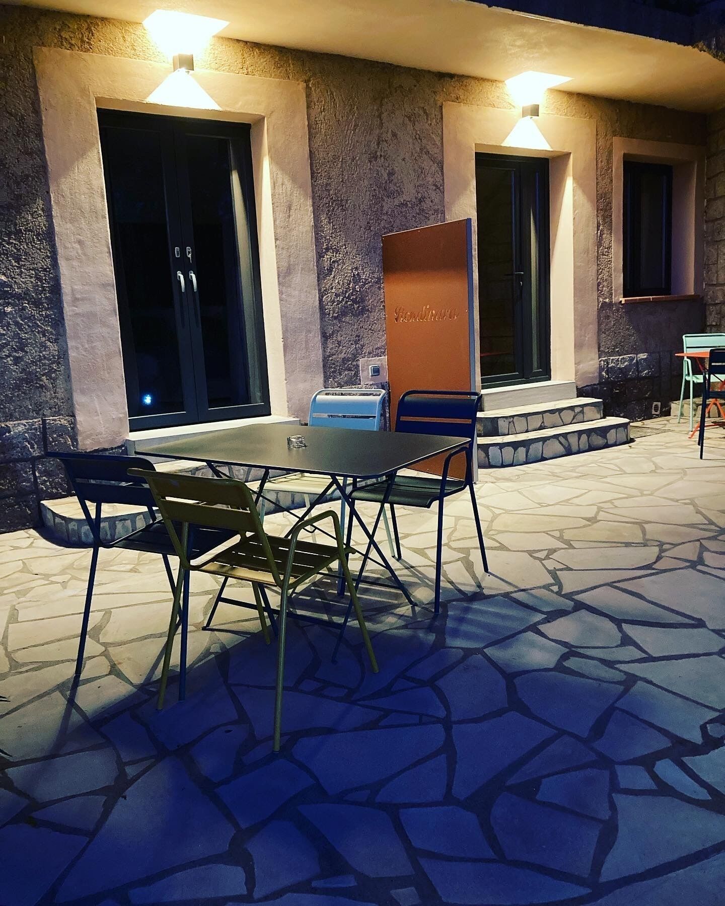 An outdoor patio with a table and chairs, a mailbox, and a building with a stone facade and two illuminated windows.