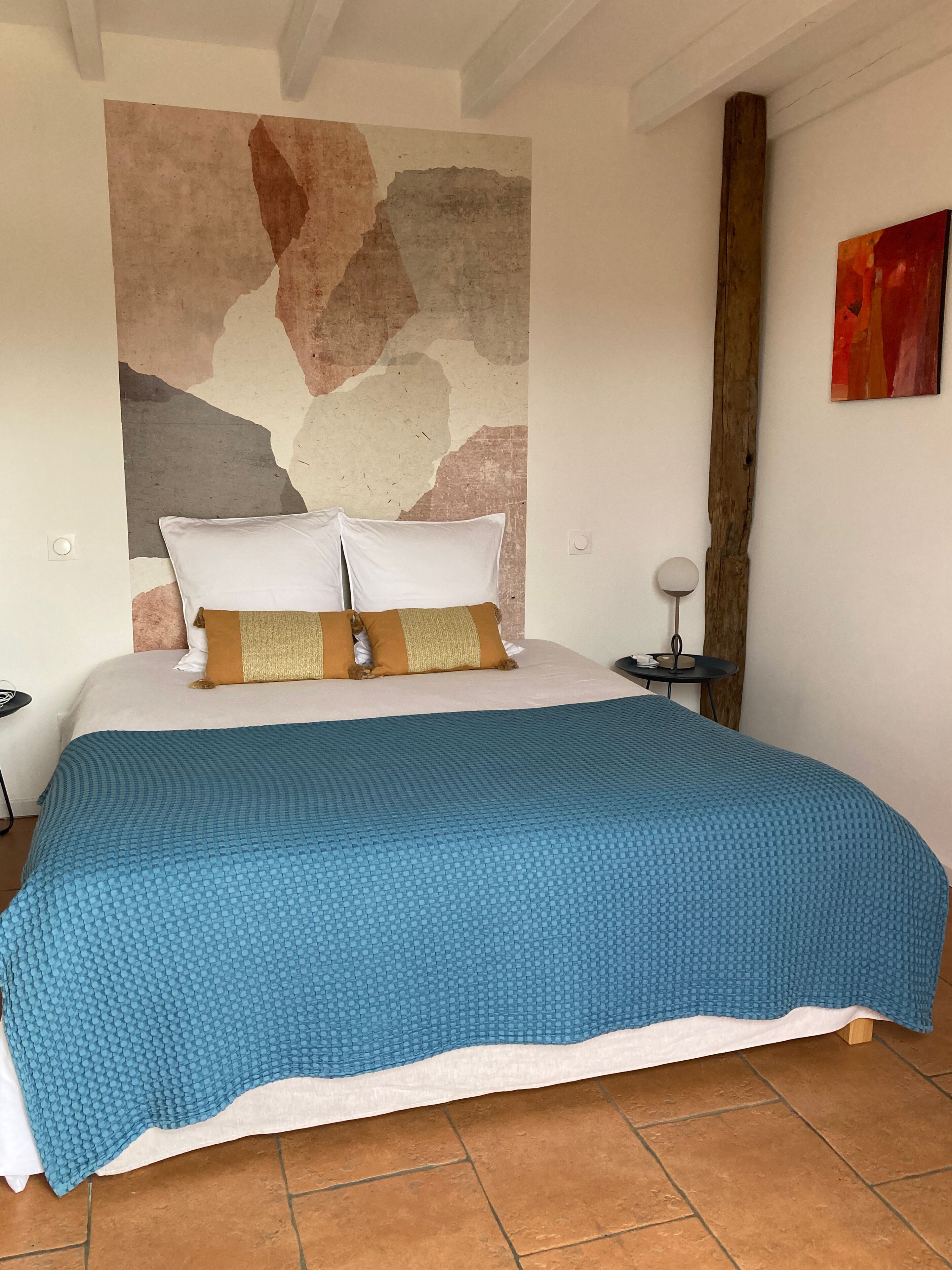 Une chambre à coucher avec un grand lit, deux oreillers, une couverture bleue, une tête de lit en bois, une table de chevet avec une lampe et un tableau abstrait rouge accroché au mur.