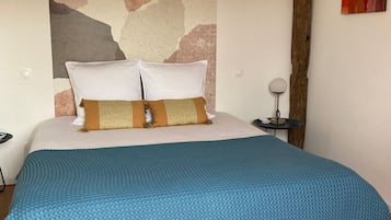 Une chambre à coucher avec un grand lit, deux oreillers, une couverture bleue, une tête de lit en bois, une table de chevet avec une lampe et un tableau abstrait rouge accroché au mur.