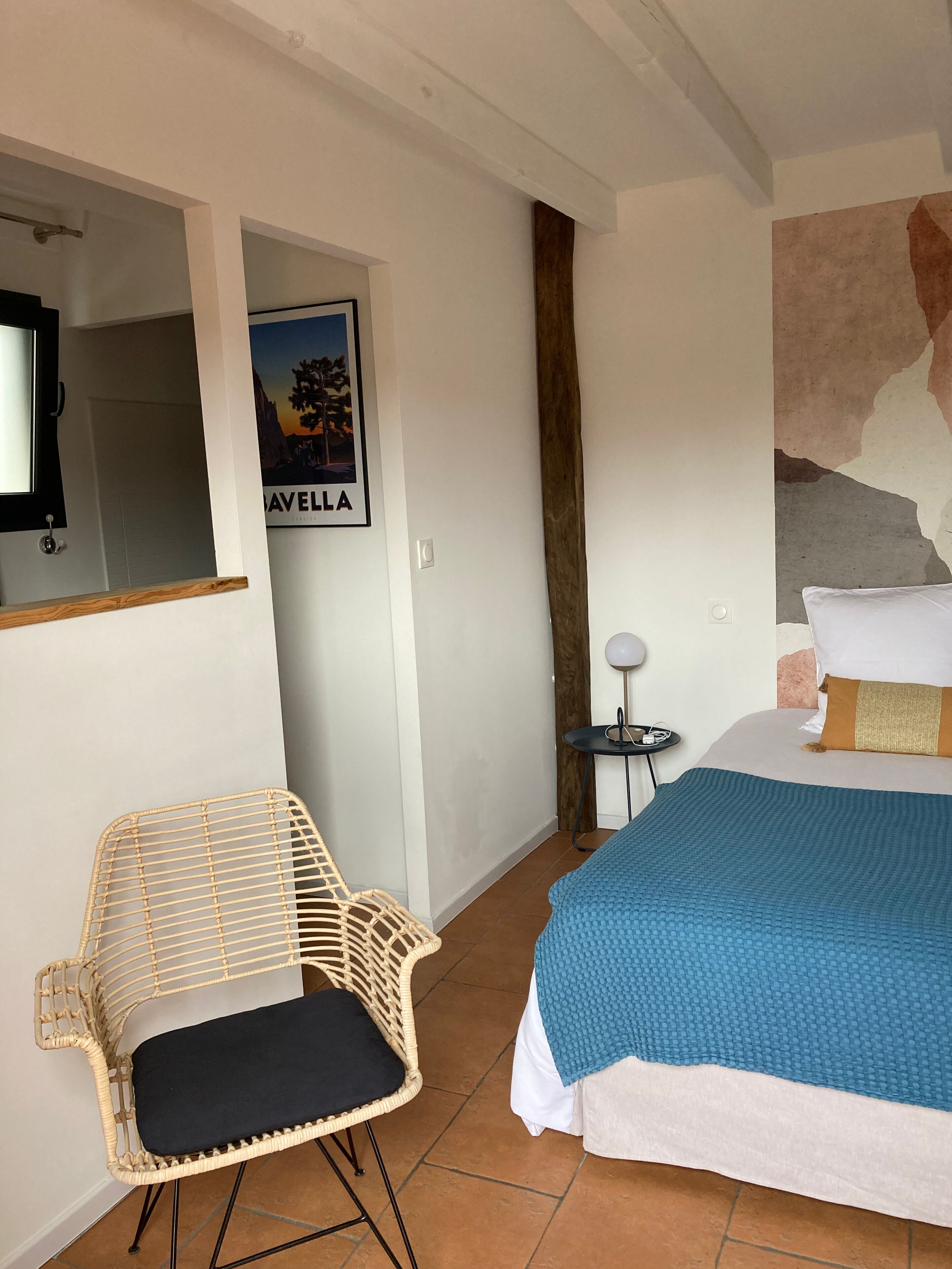 A bedroom with a bed, a wicker chair, a small table with a lamp, and a poster on the wall.
