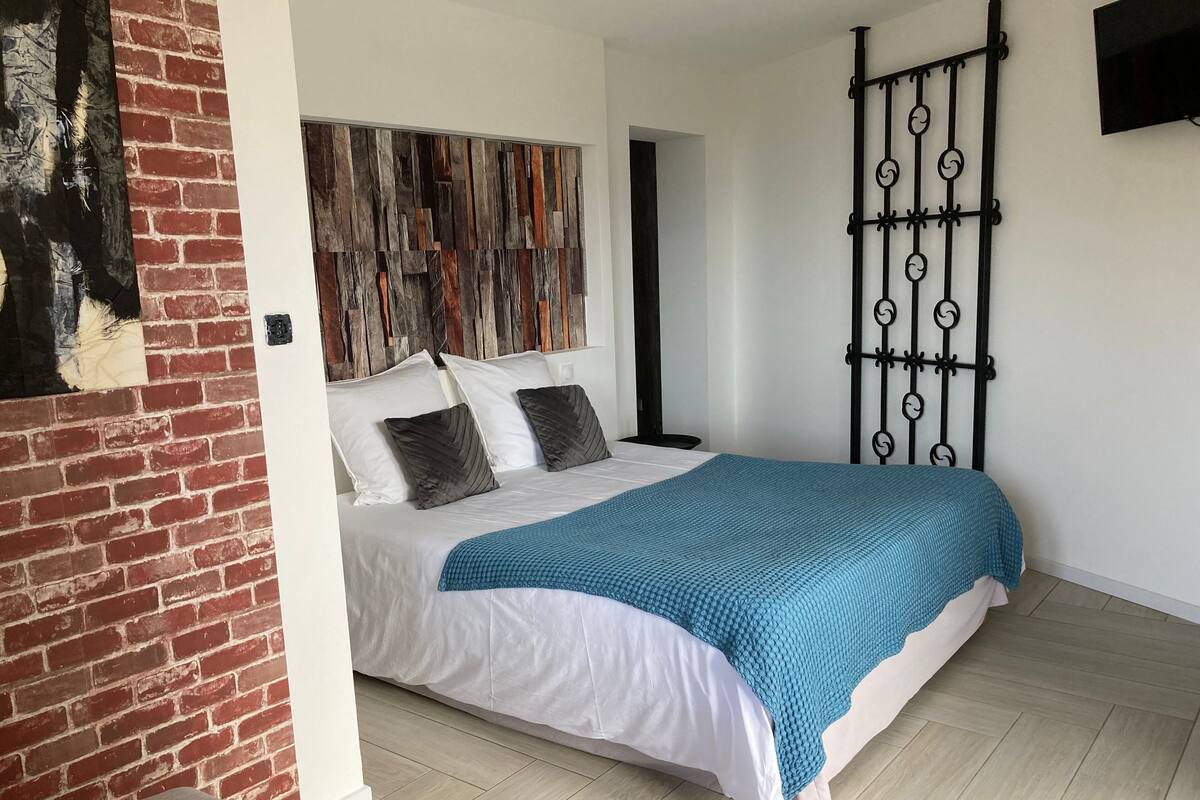 A bedroom with a bed, white bedding, a blue blanket, a brick wall, and a metal gate.