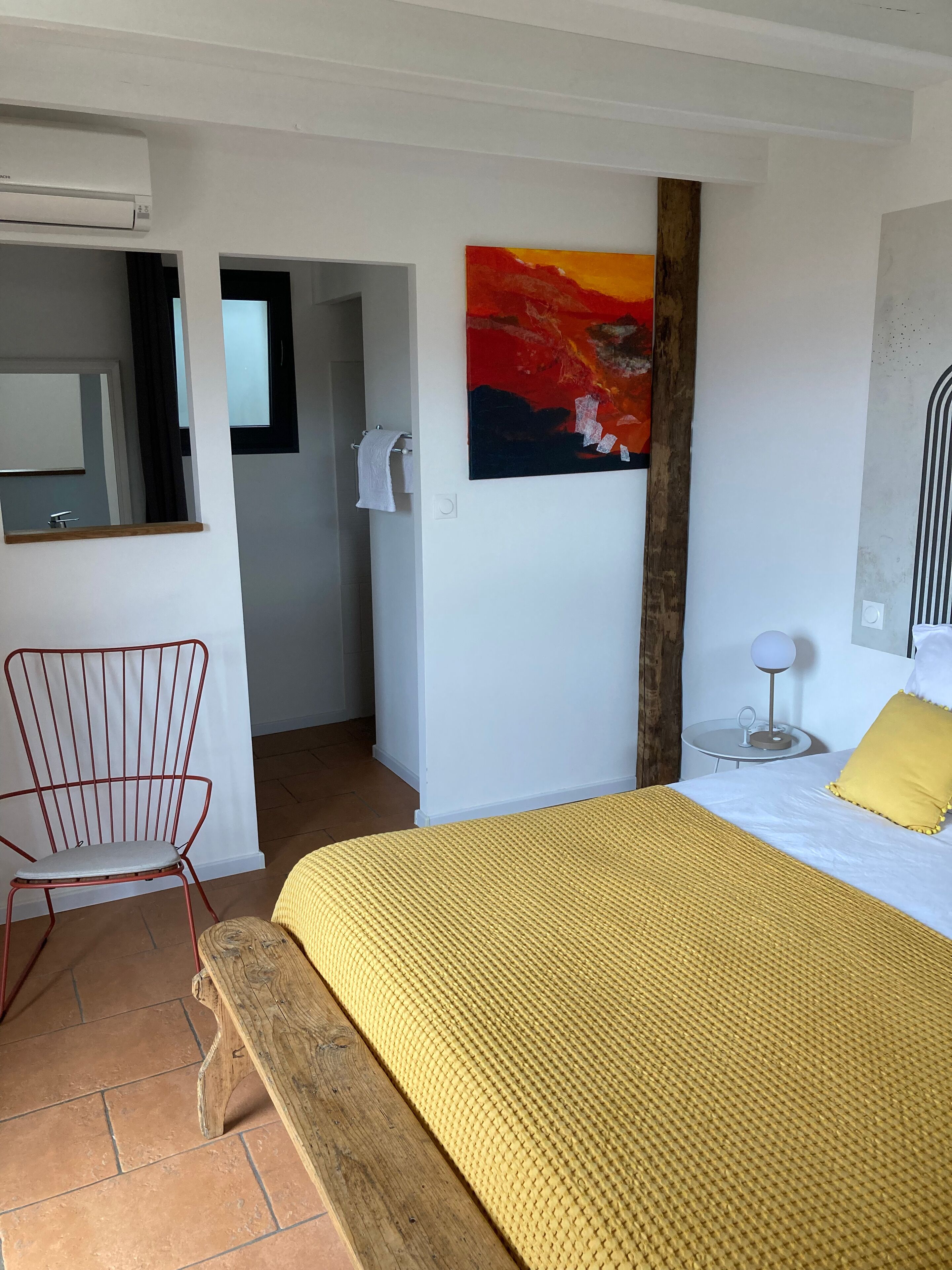 A bedroom with a bed, a chair, a painting, and a bathroom visible through an open door.
