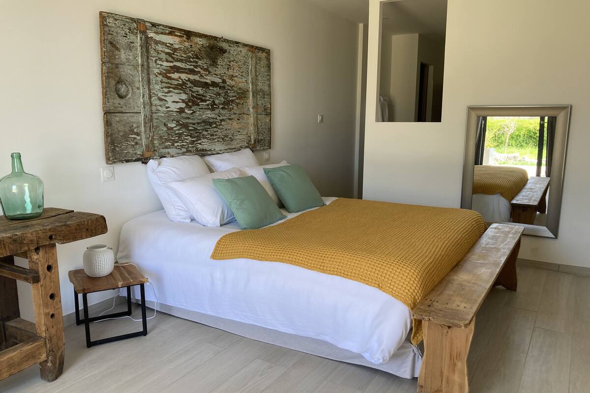 A bedroom with a bed, pillows, a wooden bench, a small table, and a large mirror.