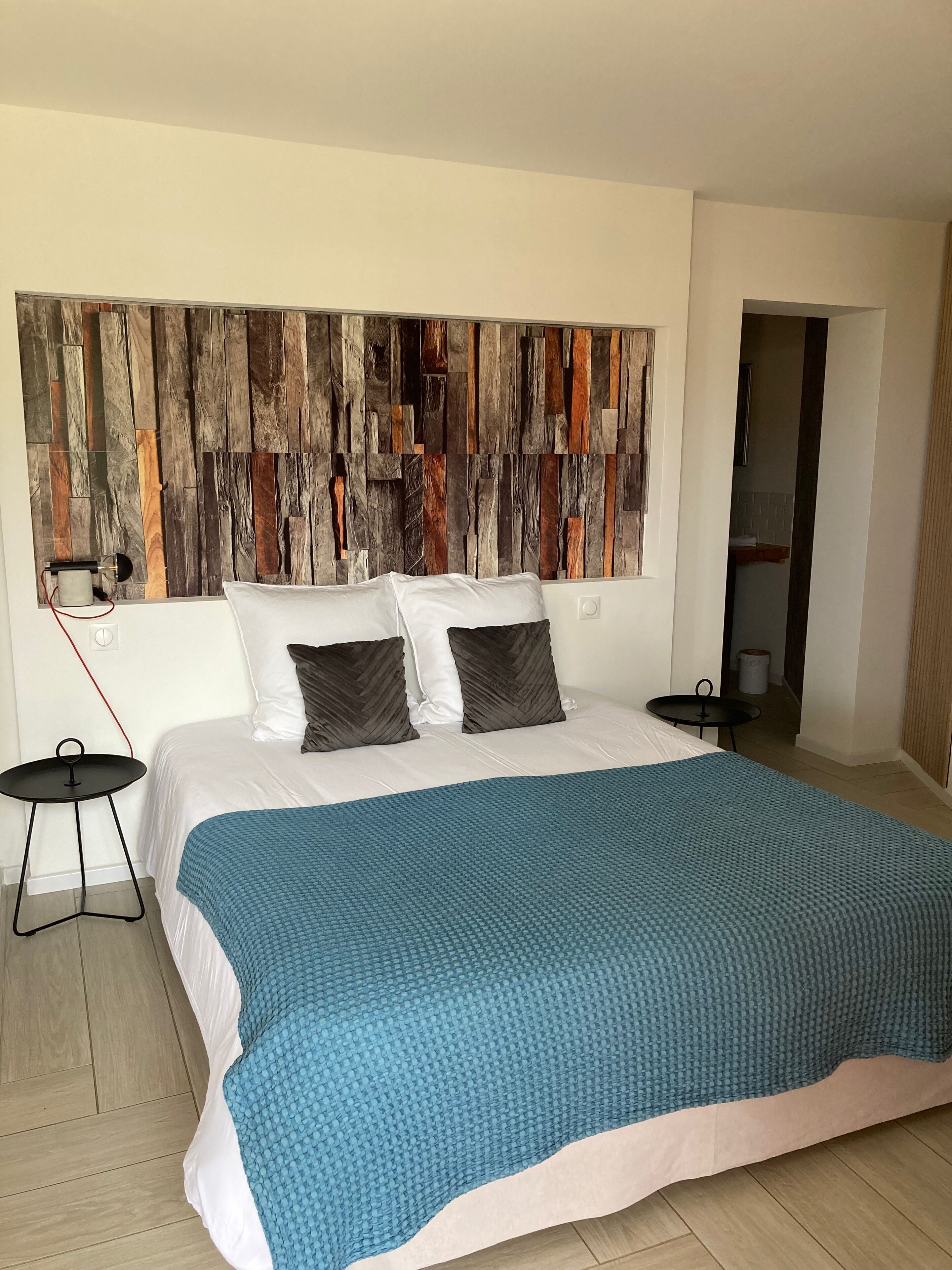 A bedroom with a large bed, two pillows, a blue blanket, a wooden wall panel, a small table, and a red power cord.