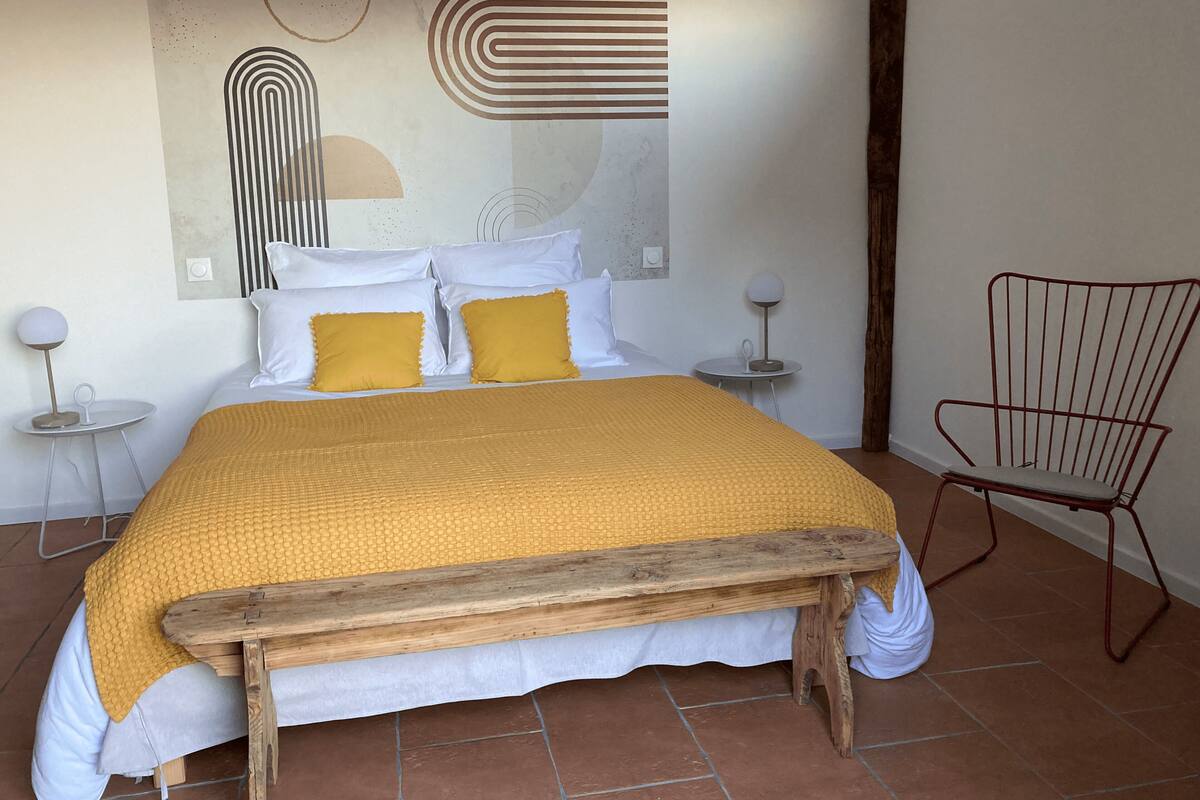 A bedroom with a wooden bed, white and yellow bedding, a red chair, and a modern abstract wall art.