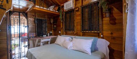 A wooden cabin room with a bed, a desk, and a window with bars.