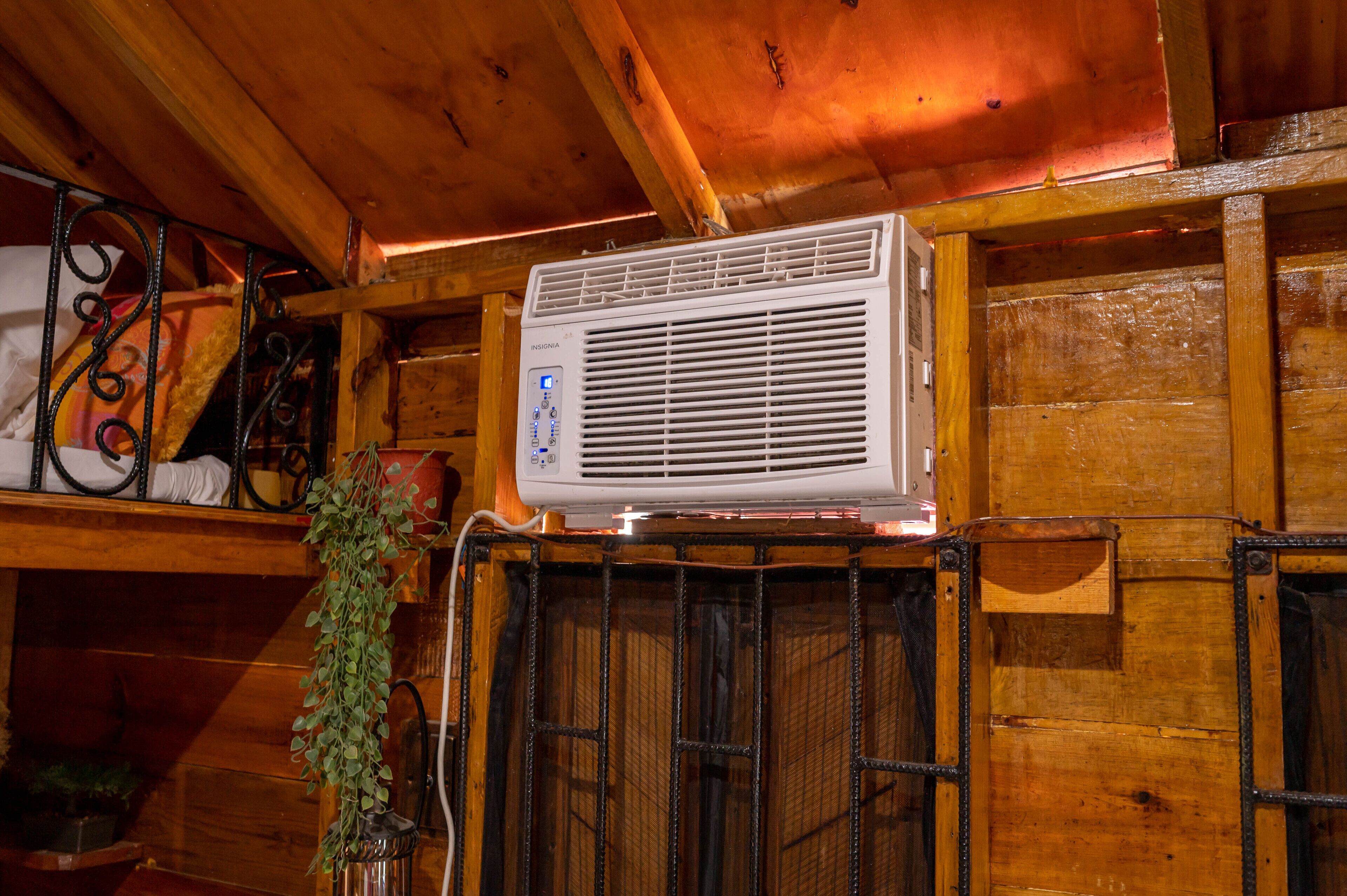 Een airconditioningunit bevestigd aan een houten wand, met een metalen rooster, een hangplant en een plank met diverse spullen.