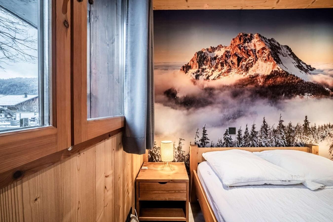 A bedroom with a wooden headboard, a bed with white bedding, a nightstand with a lamp, and a window with a view of a mountain landscape.