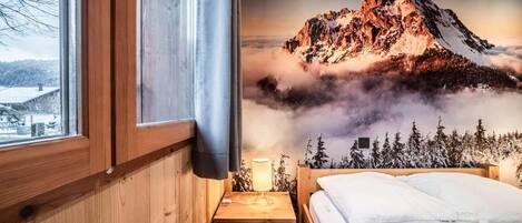 A bedroom with a wooden headboard, a bed with white bedding, a nightstand with a lamp, and a window with a view of a mountain landscape.