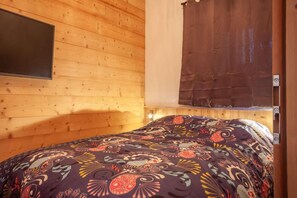 Ein Zimmer einer Holzhütte mit einem Bett, einem Wandfernseher und einem Fenster mit Vorhang.