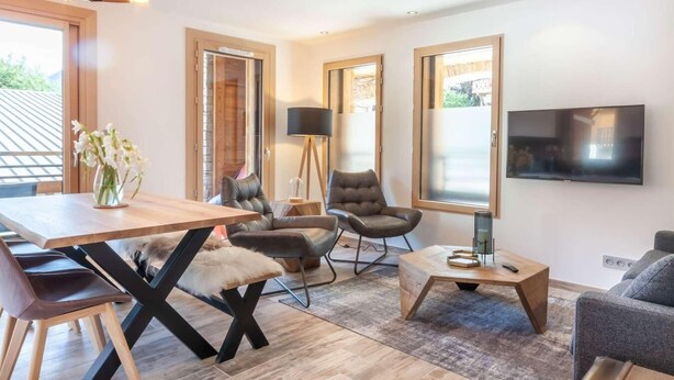 A modern living room with a wooden dining table, leather chairs, a TV mounted on the wall, and large windows.