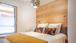 A bedroom with a wooden headboard, a large bed with white bedding and a mustard-colored throw blanket, and a geometric pendant light.