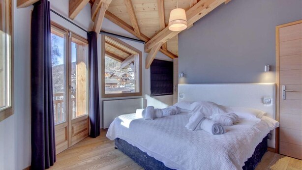 Une chambre à coucher avec un grand lit, des poutres en bois, une fenêtre avec des rideaux et une vue sur un village.