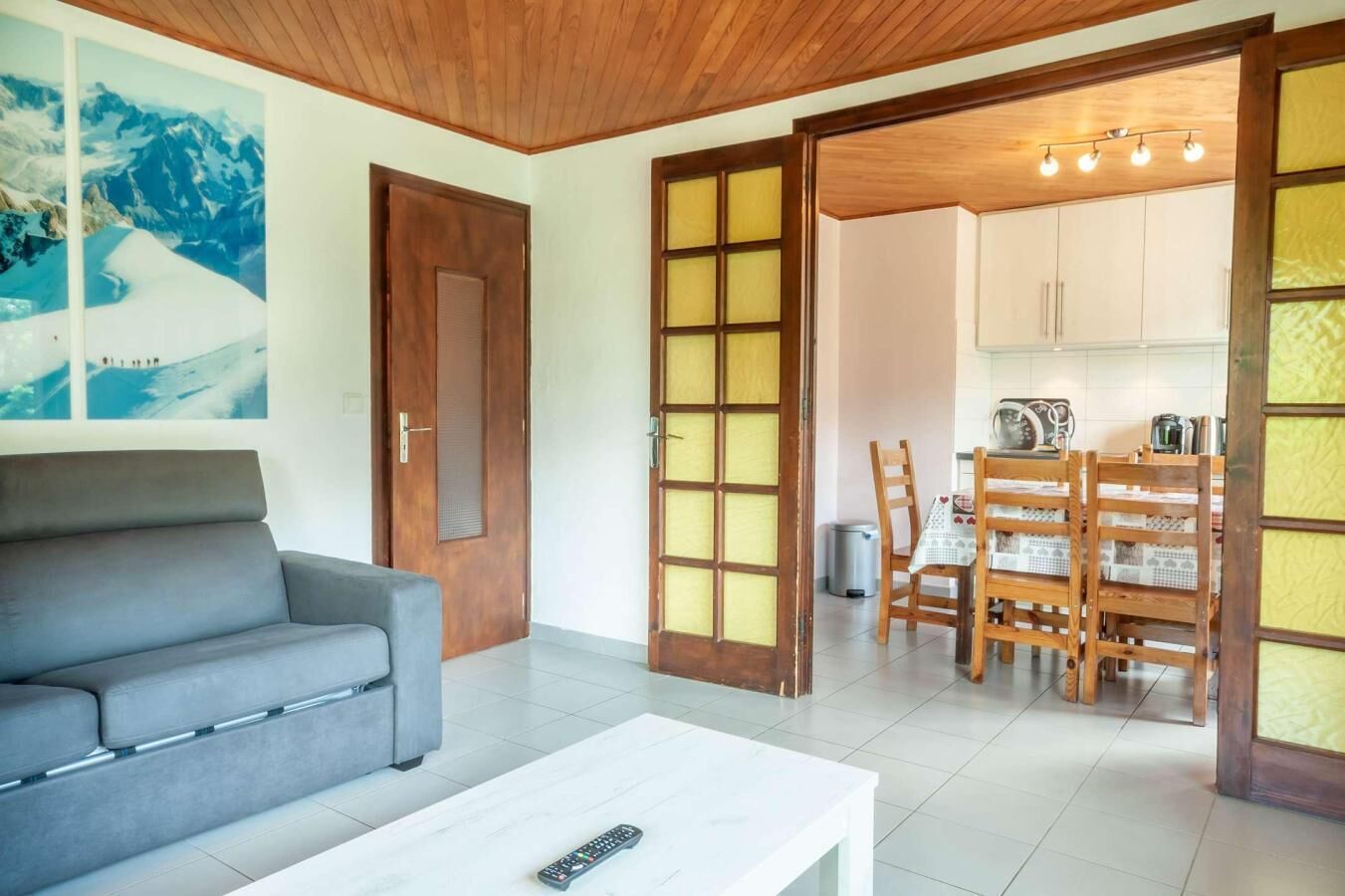 A living room with a grey sofa, a white coffee table, and a wooden door leading to a dining area with a table and chairs.