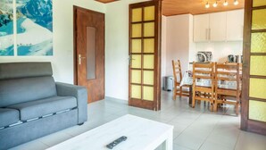 Una sala de estar con un sofá gris, una mesa de centro blanca y una puerta de madera que da a un comedor con una mesa y sillas.