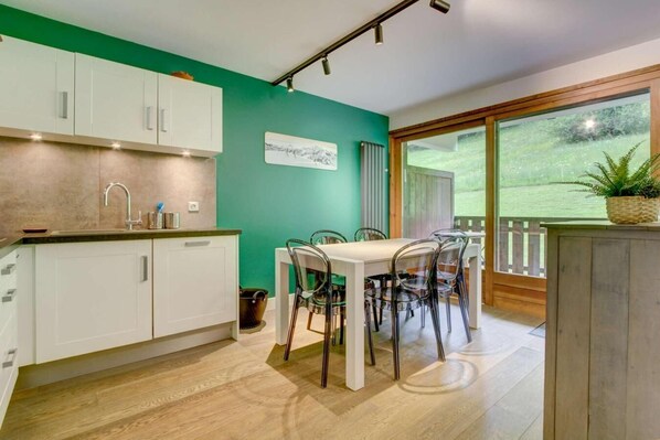 A modern kitchen with white cabinets, a dining table with chairs, and a view of a garden through large windows.