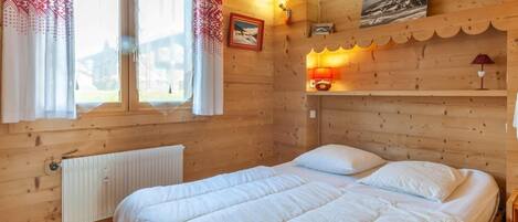 Une chambre dans un chalet en bois, avec un lit, une fenêtre avec des rideaux, un radiateur et des lampes fixées au mur.