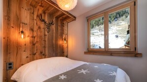A bedroom with a wooden headboard, a bed with a white comforter, a gray blanket with snowflake patterns, and a window with a view of a snowy landscape. - 50m from ski slopes, 1st floor, swimming pool, sauna, hammam, balcony, fireplace or stove, tv (Champagny-en-Vanoise)
