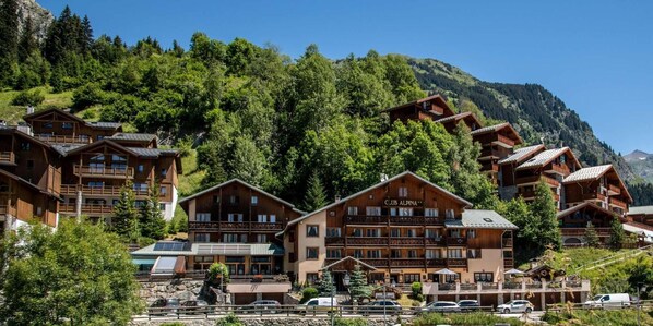 Un conjunto de edificios de madera con balcones, identificados como "CLUB ALPINA", frente a un paisaje de colinas y montañas verdes.