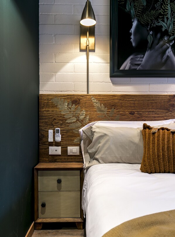 A bedroom with a wooden headboard, a bedside table with a phone and a power outlet, a wall-mounted lamp, and a bed with white bedding and a brown throw blanket. - Hotel Celestino Boutique (Medellín)
