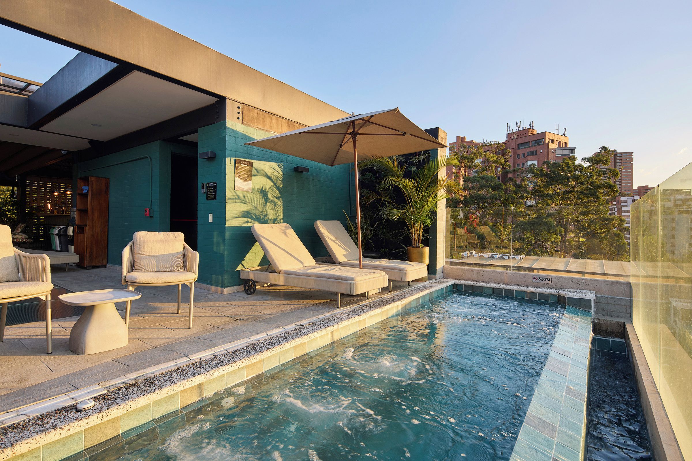A rooftop pool area with lounge chairs and a table, surrounded by a modern building with a blue wall and a glass enclosure.