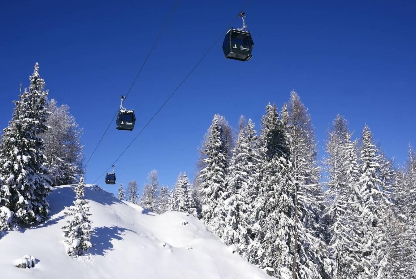 Un télésiège avec des cabines qui montent une pente enneigée, entouré de grands arbres chargés de neige, sous un ciel bleu dégagé.