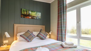 A neatly made bed with a colorful plaid blanket and pillows, a painting of a forest on the wall, and a window with curtains. - Cedar Lodge (Newark)