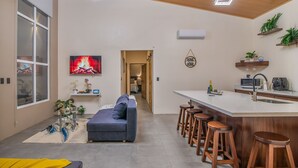 Ein modernes Wohnzimmer mit Sofa, einem Fernseher, der einen Kamin zeigt, und einer Küchenzeile mit Bar und Hockern.
