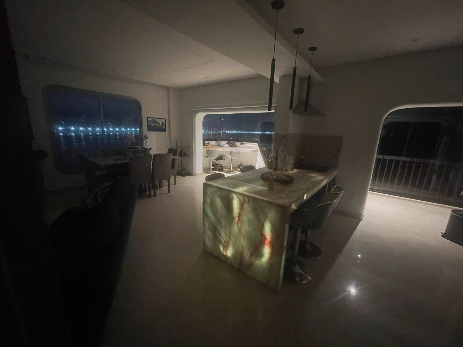Una cocina moderna con una isla central, zona de comedor y ventanas amplias con vistas a un paisaje urbano nocturno.