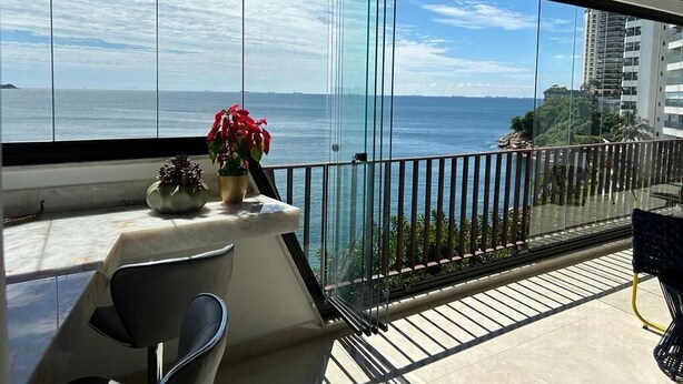 A balcony with a glass railing overlooking the sea, a table, and two chairs. - Apartamento Astúrias (Guarujá)