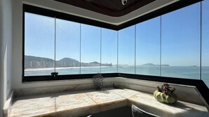 A corner room with a large window overlooking a beach and cityscape. - Apartamento Astúrias (Guarujá)