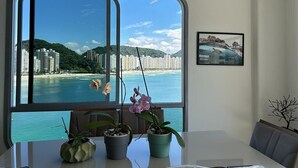 A room with a large window offering a view of a beach and cityscape. There are potted plants on the windowsill. - Apartamento Astúrias (Guarujá)