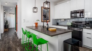 A modern kitchen with white cabinets, a stainless steel oven, and a built-in microwave. There are two hanging pendant lights and a small island with stools. - Pura Vida House - 2 King Bed - Entire Home (Sacramento)