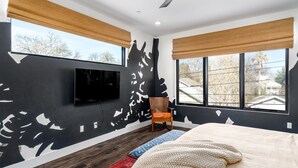 A bedroom with a bed, a television mounted on the wall, a chair, and large windows with blinds. - Pura Vida House - 2 King Bed - Entire Home (Sacramento)