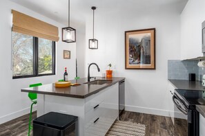 Une cuisine moderne dotée d’un îlot central, d’un évier et d’un lave-vaisselle. On y trouve une fenêtre avec un store, une chaise verte et un tableau encadré accroché au mur.