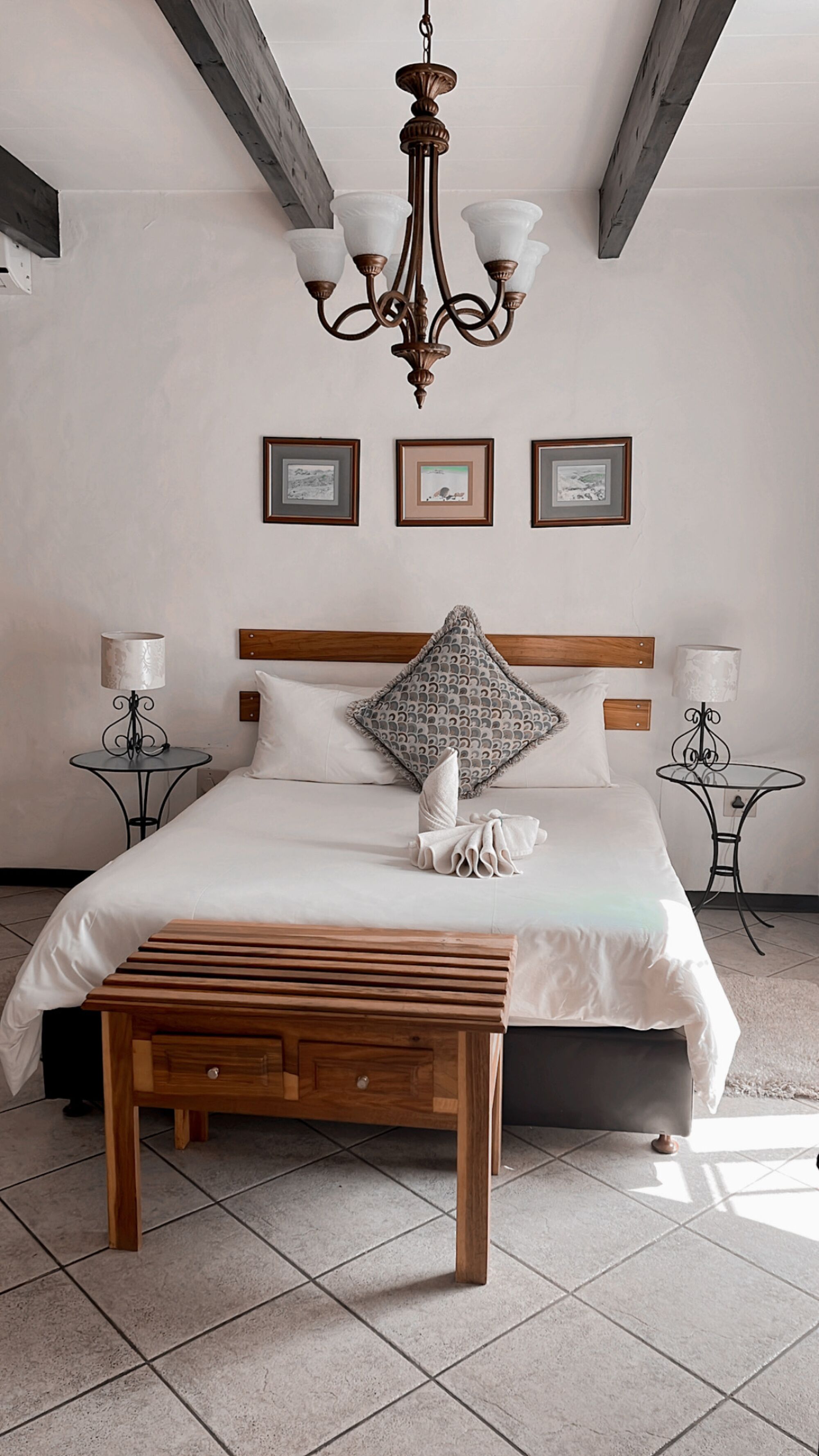 A bedroom with a large bed, wooden bench, chandelier, framed pictures, and two bedside tables with lamps.