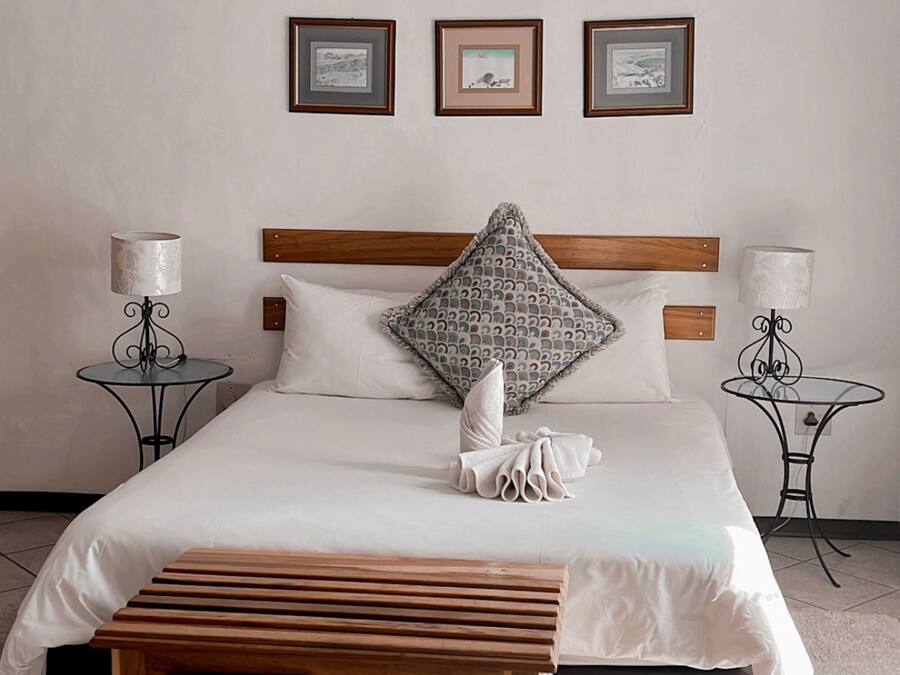 A bedroom with a large bed, wooden bench, chandelier, framed pictures, and two bedside tables with lamps.