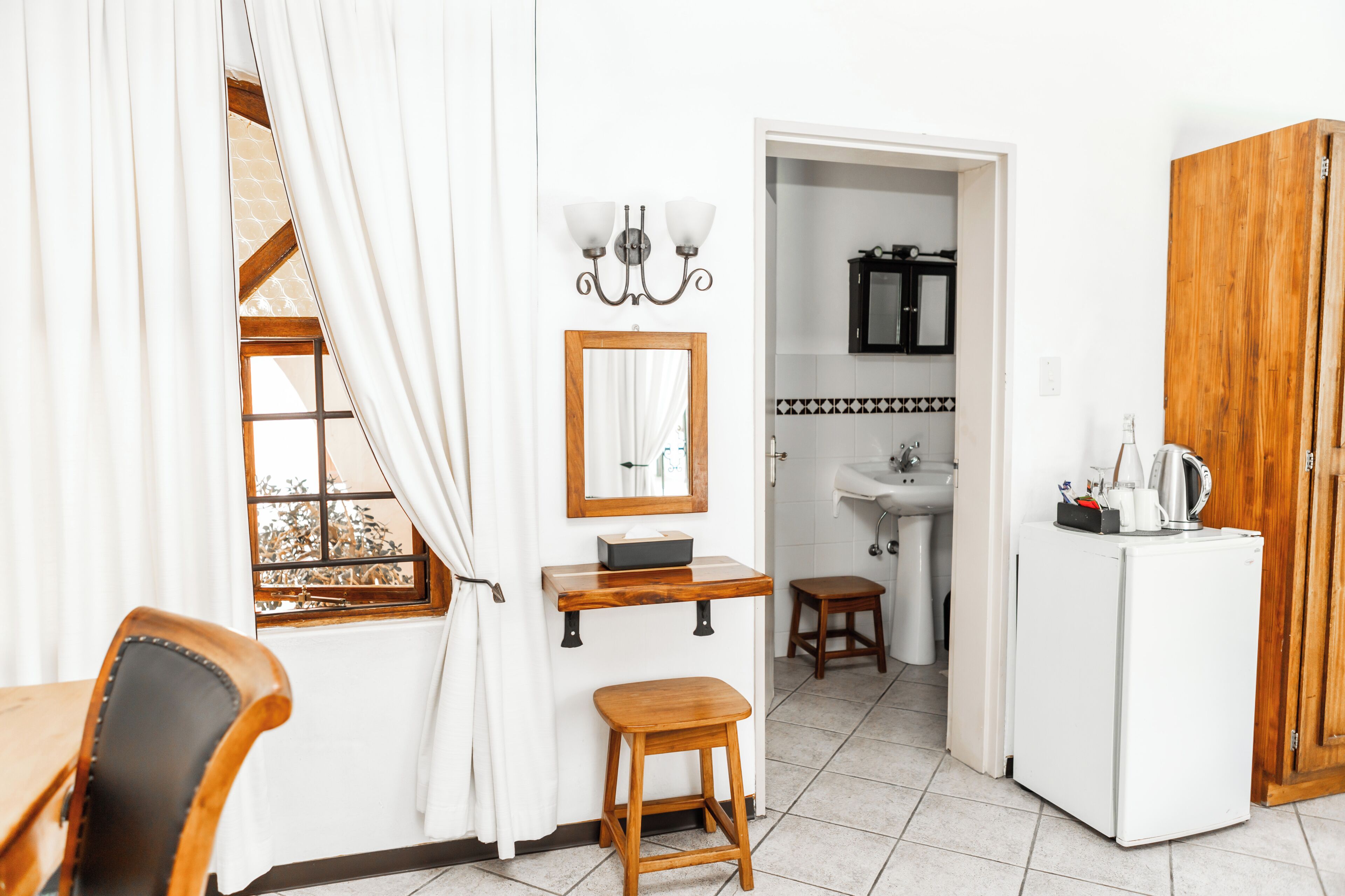 Ein Zimmer mit einem Holzstuhl, einem kleinen Hocker, einem weißen Kühlschrank, einem Holzboden mit Spiegel und Kiste sowie einem Blick durch ein Fenster mit weißen Vorhängen.
