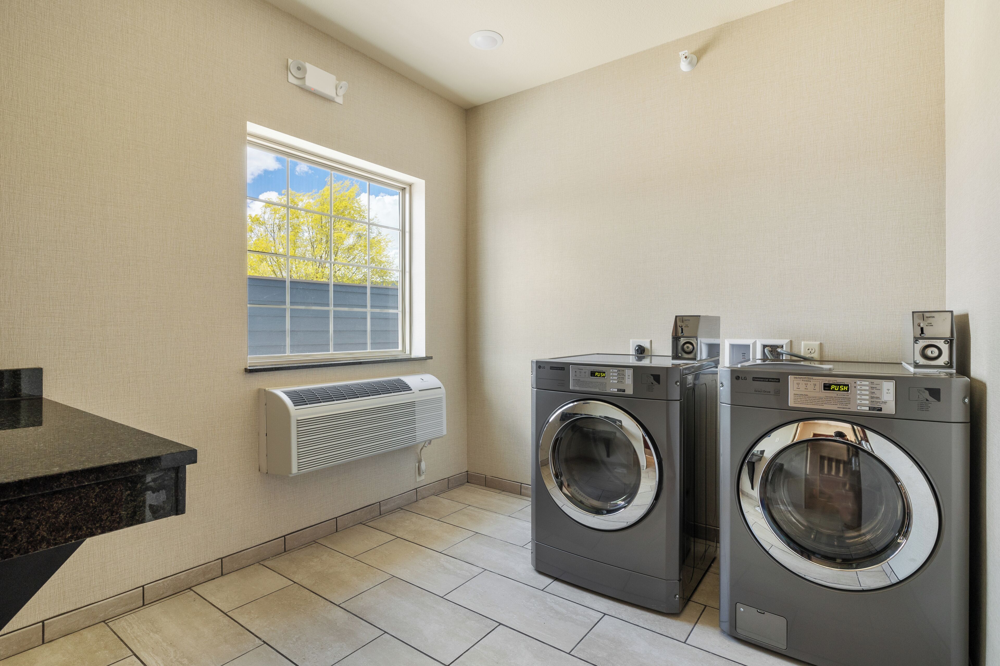 A laundry room with two front-loading washing machines, a built-in air conditioner, and a window with a view of trees.