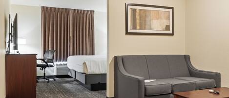 A hotel room with a bed, a gray sofa, a wooden table, a desk with a chair, and a framed picture on the wall.