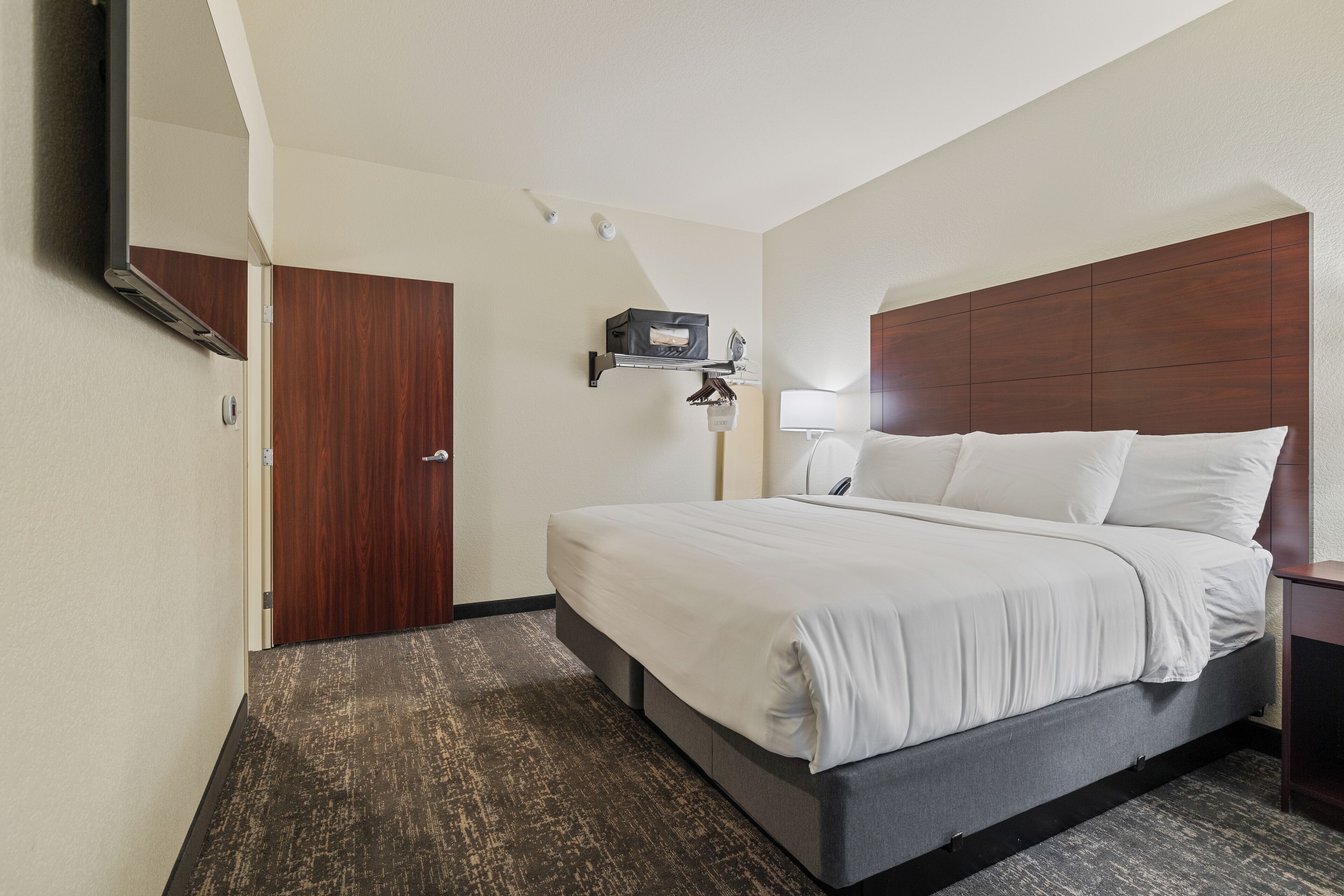 Habitación de hotel con cama, mesita de noche, lámpara, estante en la pared y una puerta.