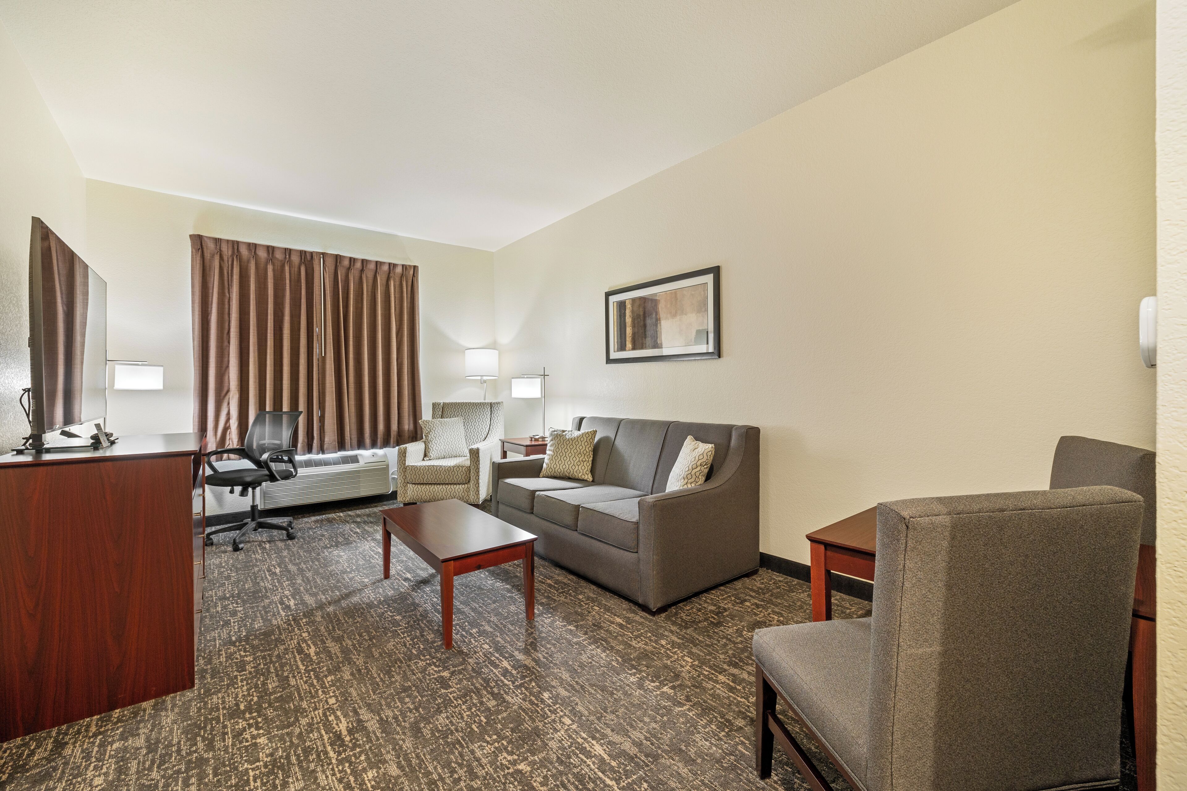 A hotel room with a wooden desk, a grey sofa, a grey armchair, a small table, and a large window with curtains.