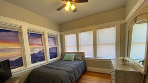 Un dormitorio con cama, piso de madera, ventilador de techo y ventanas grandes con persianas.