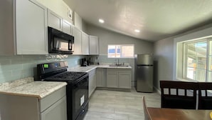 Cocina compacta con estufa, microondas y refrigerador, armarios color gris claro y ventana que proporciona luz natural.